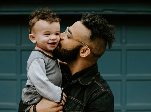 Father joyfully kissing his smiling toddler, both wearing casual clothing, symbolizing family support and connection in early childhood development.