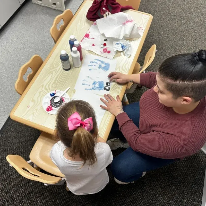 Child engaged in creative art activity with ABA therapist, using paint and paper at a small table in a supportive learning environment.