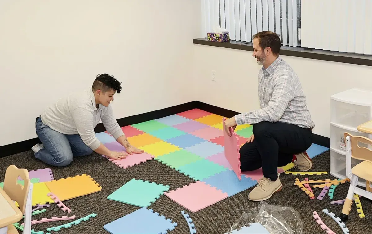 Two individuals arranging colorful interlocking foam mats in a developmental services setting, emphasizing teamwork and a supportive environment for children.