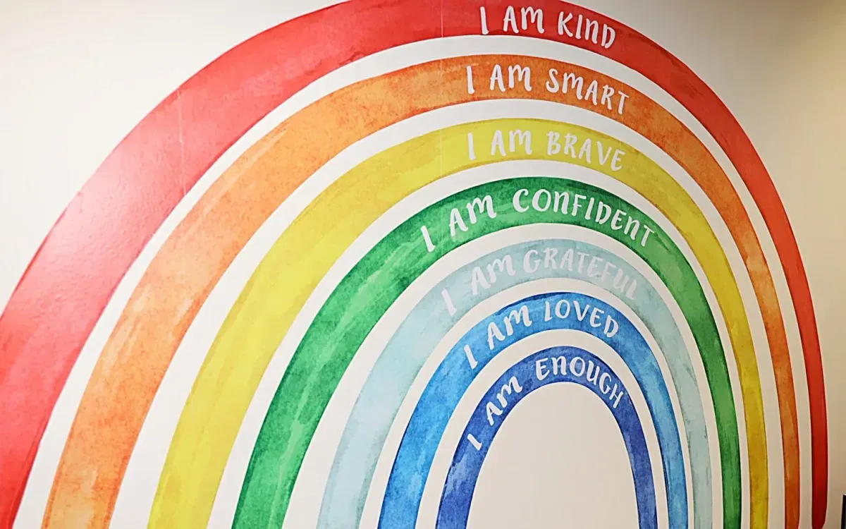 Colorful rainbow mural with affirmations: &ldquo;I AM KIND,&rdquo; &ldquo;I AM SMART,&rdquo; &ldquo;I AM BRAVE,&rdquo; &ldquo;I AM CONFIDENT,&rdquo; &ldquo;I AM GRATEFUL,&rdquo; &ldquo;I AM LOVED,&rdquo; &ldquo;I AM ENOUGH,&rdquo; reflecting a supportive environment at First Steps Developmental Services.