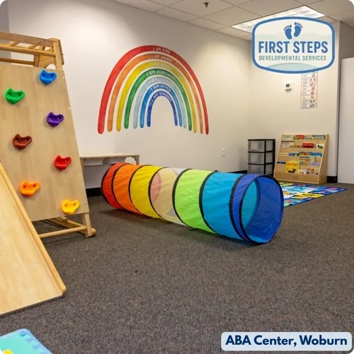 Brightly colored play area featuring a rainbow mural, climbing wall with grips, and a tunnel, designed for engaging and supportive ABA therapy at First Steps Developmental Services, Woburn.
