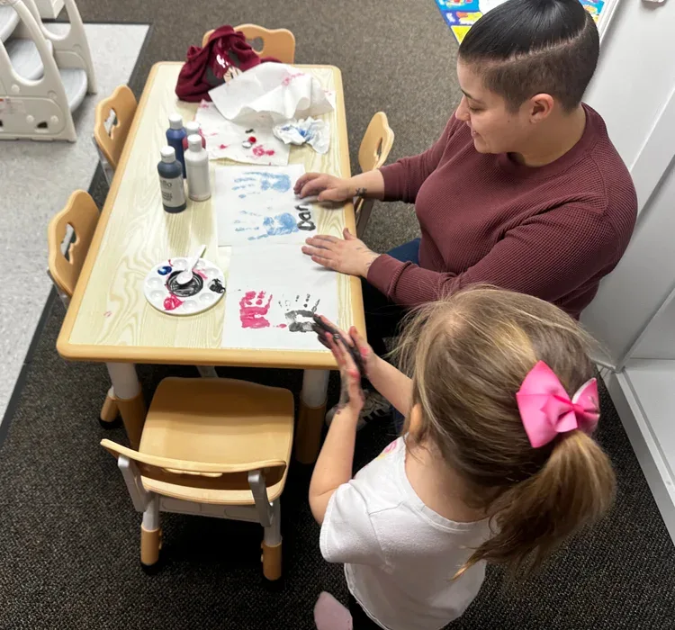 Child and caregiver engaged in creative art activity at a table, using paint and making handprints, emphasizing child-led play-based learning in a nurturing environment at First Steps Developmental Services