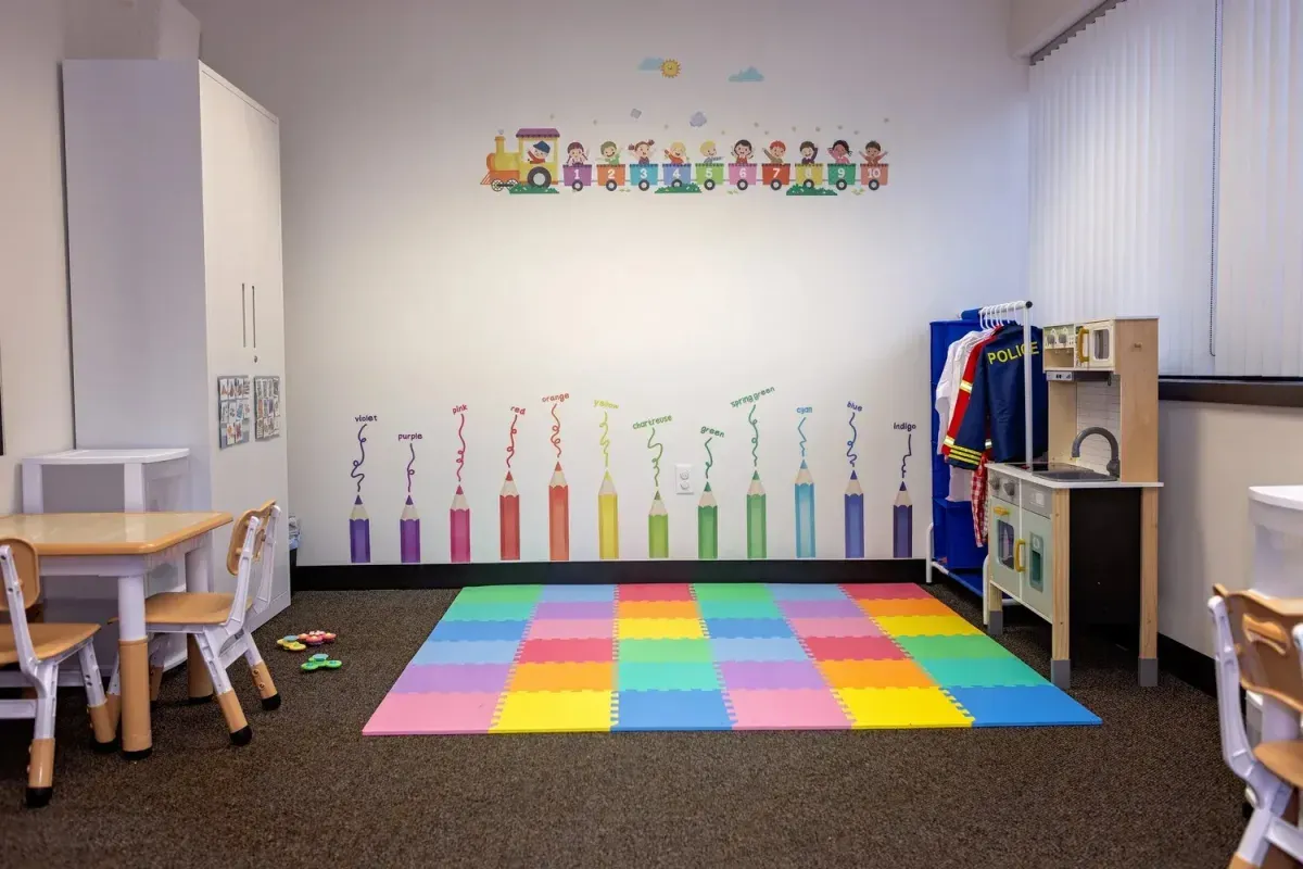 Colorful playroom with interactive learning elements, featuring a train mural, vibrant pencil graphics labeled with colors, a soft play mat, and a play kitchen setup, supporting child-led play and joyful learning at First Steps ABA Therapy Center