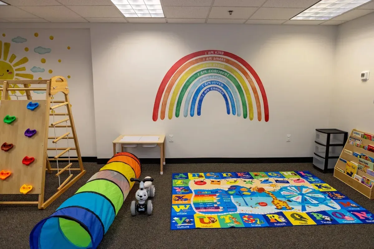 Colorful playroom with climbing structure, tunnel, educational rug, and motivational rainbow mural featuring affirmations for children, emphasizing child-led learning and exploration at First Steps ABA Therapy Center