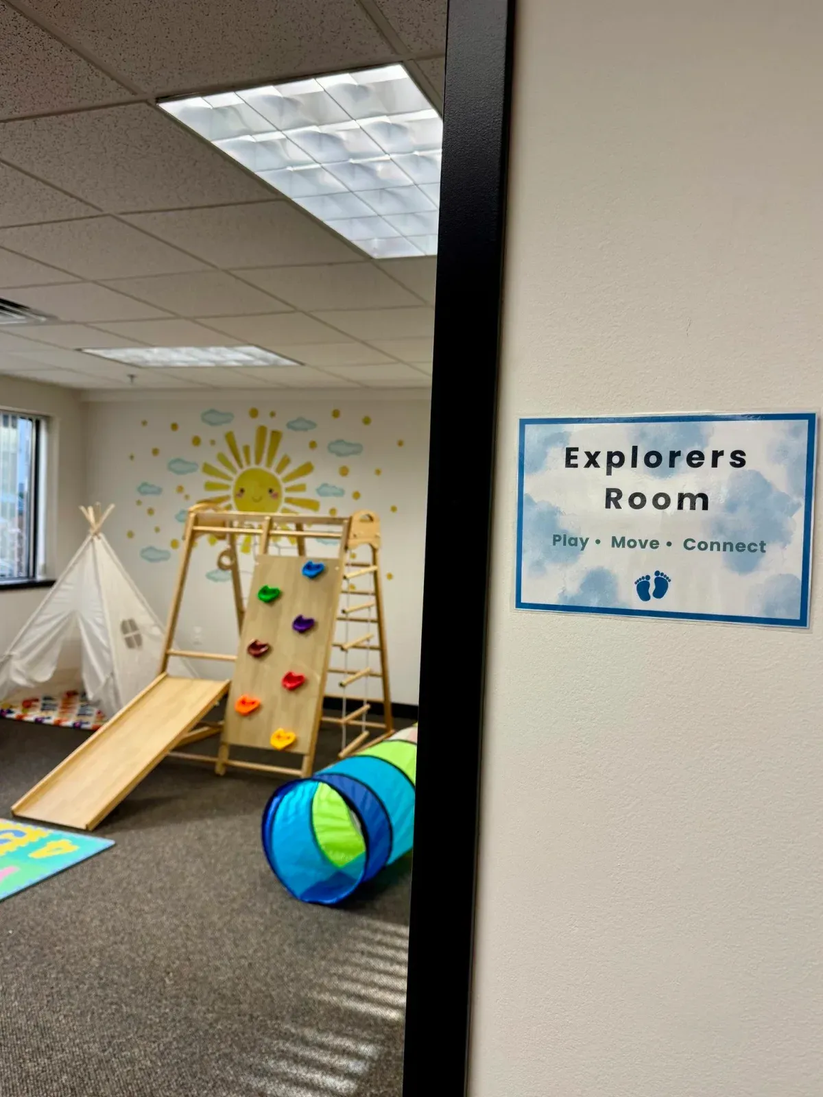 Explorers Room at First Steps Developmental Services featuring a climbing wall, slide, play tunnel, and cheerful sun mural, emphasizing child-led play and exploration